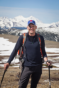 Ryan stands with his walking sticks, wearing his backpack and No Barriers Warriors hat with snowy mountains in the background
